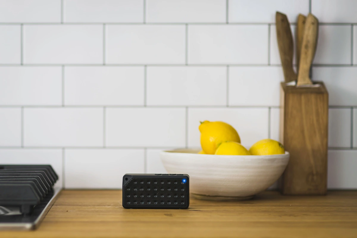 Altavoz Bluetooth portátil encendido sobre una encimera de cocina de madera, junto a un cuenco de limones