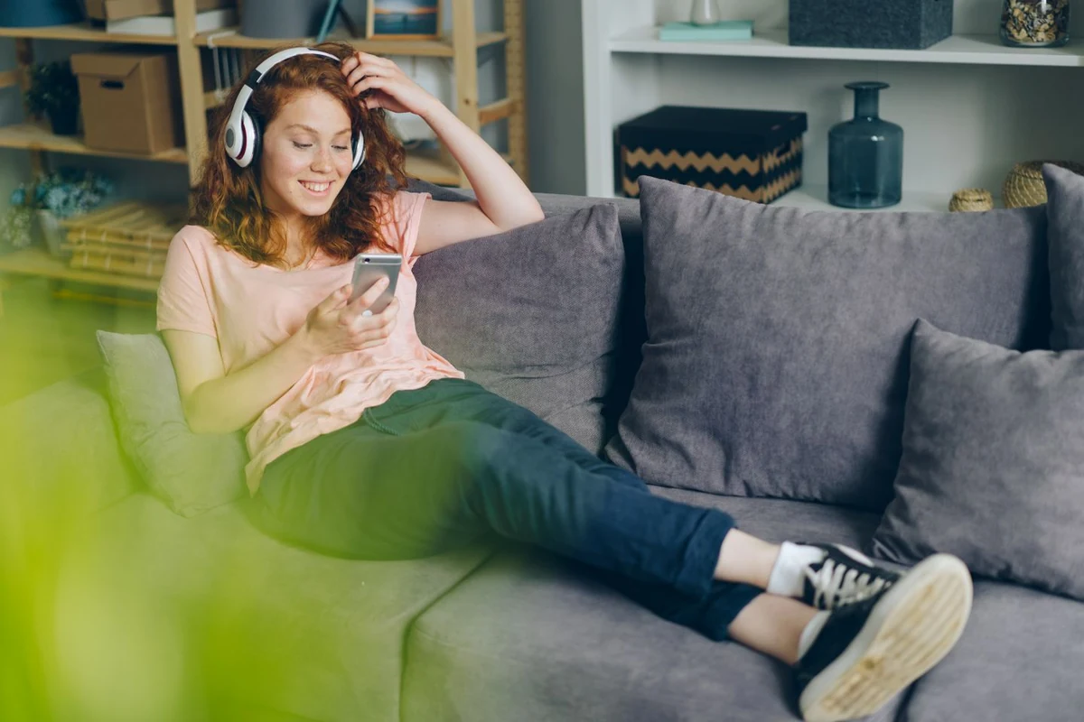 Persona escuchando música con auriculares de diadema y el móvil en la mano, sentada en el sofá de un salón