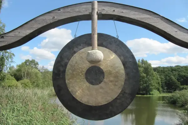 Gong de bronce bicolor colgado de un soporte de madera, con el mazo de fieltro apoyado en el disco y un lago al fondo