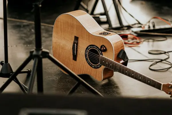 Guitarra electroacústica con cutaway y el panel del previo a la vista, apoyada en un escenario junto a pies de micro y cables